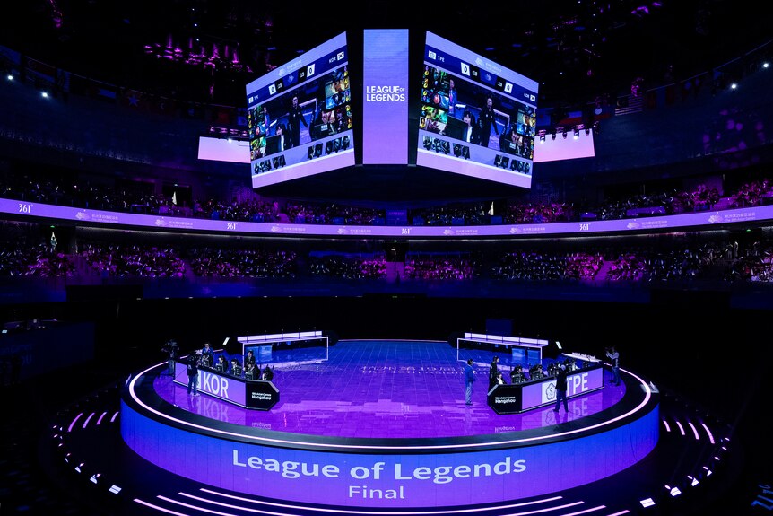 A wide shot of two esports teams behind computers on a circular stage with a large scoreboard above them.