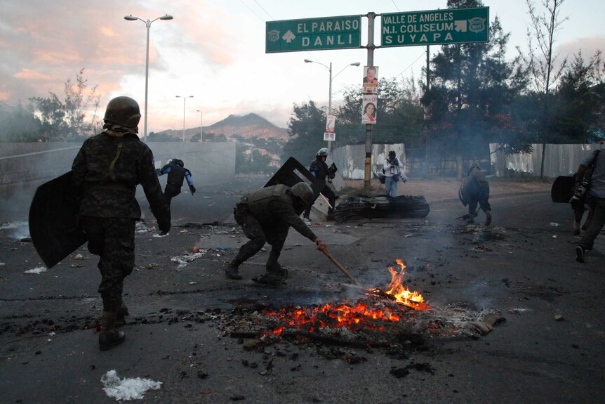 A group of armoured police walk along a road and clear debris.