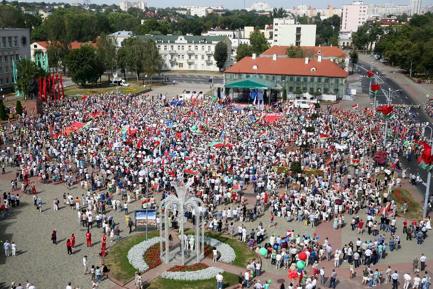 A crowd of hundreds, many wearing white, wave flags as they stand in a sunny square and listen to a speaker on a raised stage