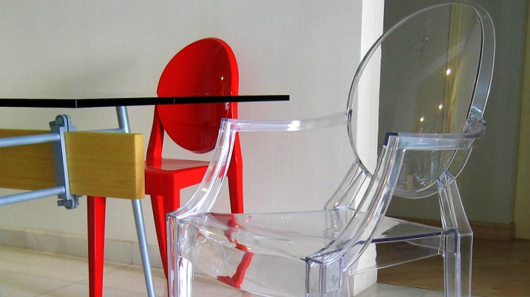 You view a transparent Louis Ghost and bright red Victoria Ghost chairs next to a table of glass and steel.