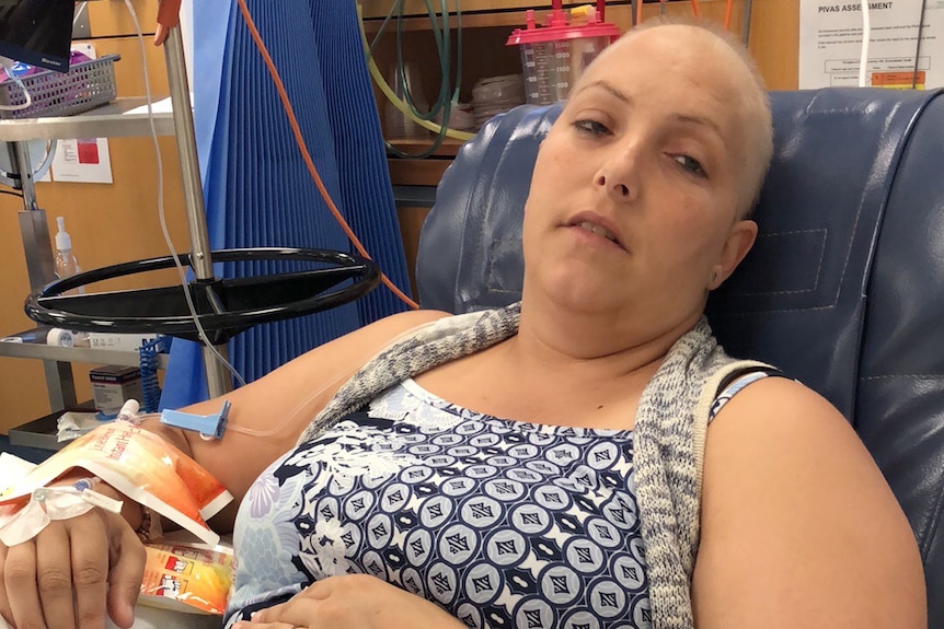 A woman who is bald and undergoing treatment for cancer lies on a bed with a tube in her arm.