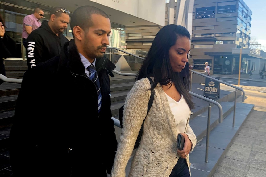 A woman in sunglasses and a long cardigan walks down steps outside the District Court holding the hand of a man in a black coat.