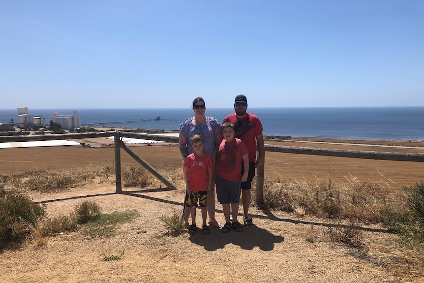 Kylie Hicklin stands with her husband and two young sons at a lookout with paddocks and the sea behind them