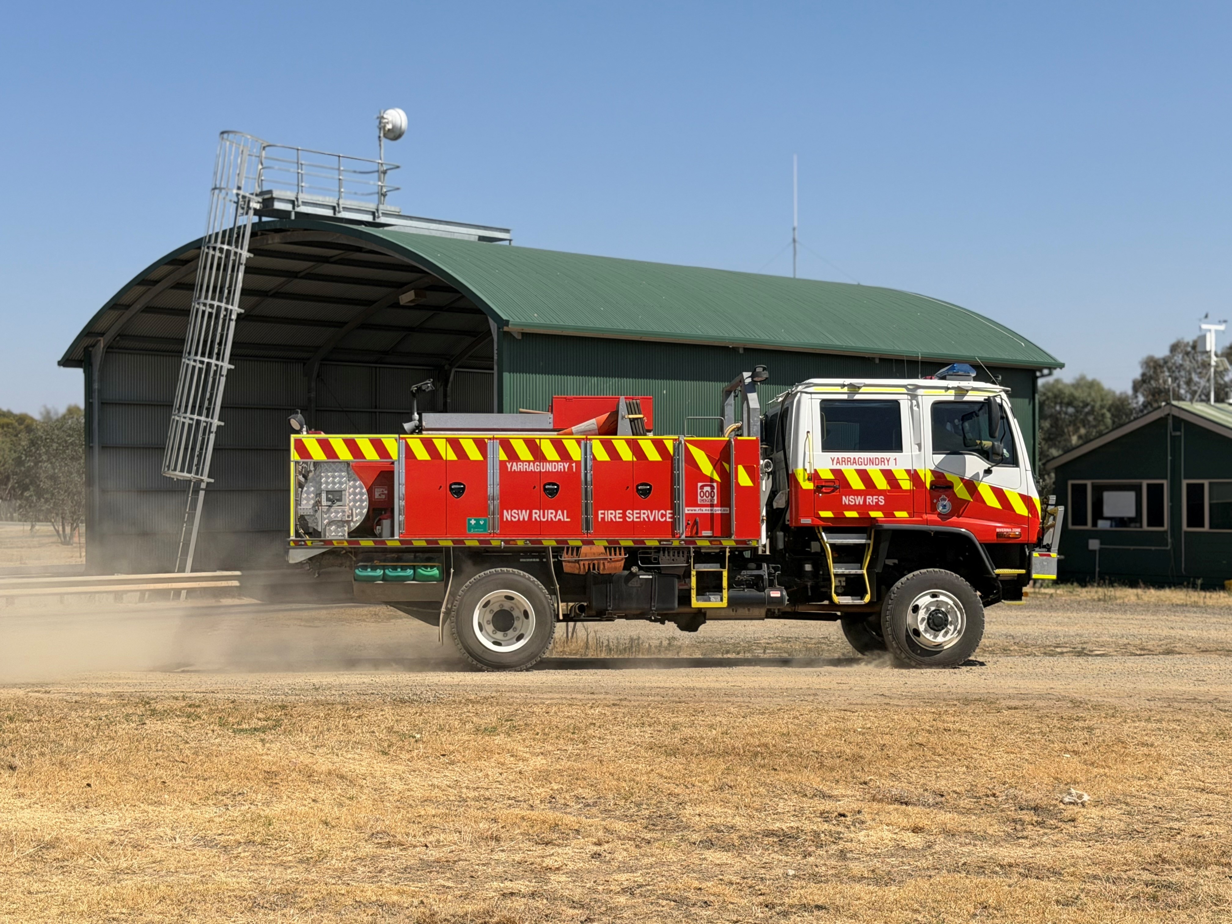 Um caminhão de bombeiros vermelho passa por uma estrada de cascalho com um grande galpão verde ao fundo.