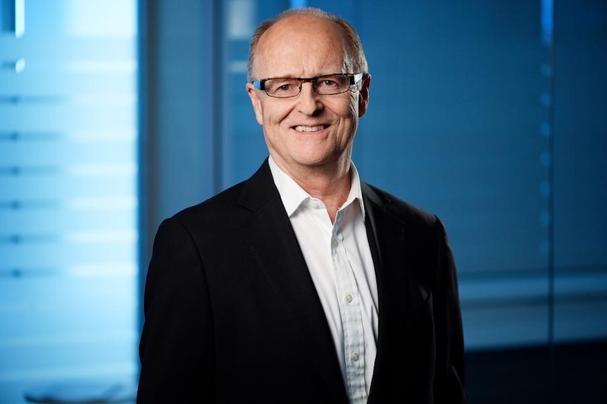 A mature man in a black jacket and white shirt with glasses smiles at the camera.