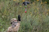 A soldier raises their hand near a small commercial drone holding an explosive. 