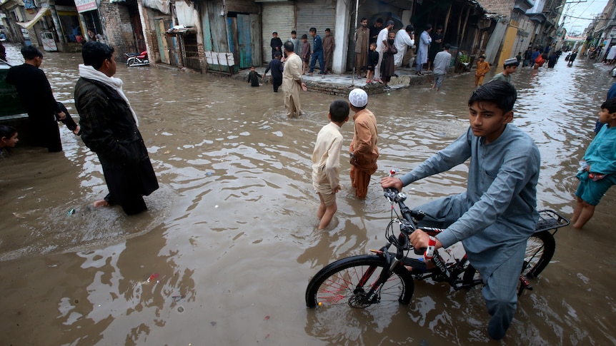 A child on a bike wades through a flooded street as people huddle under shelter looking on