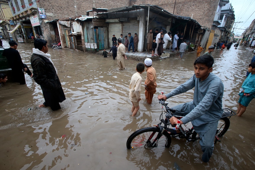 A child on a bike wades through a flooded street as people huddle under shelter looking on