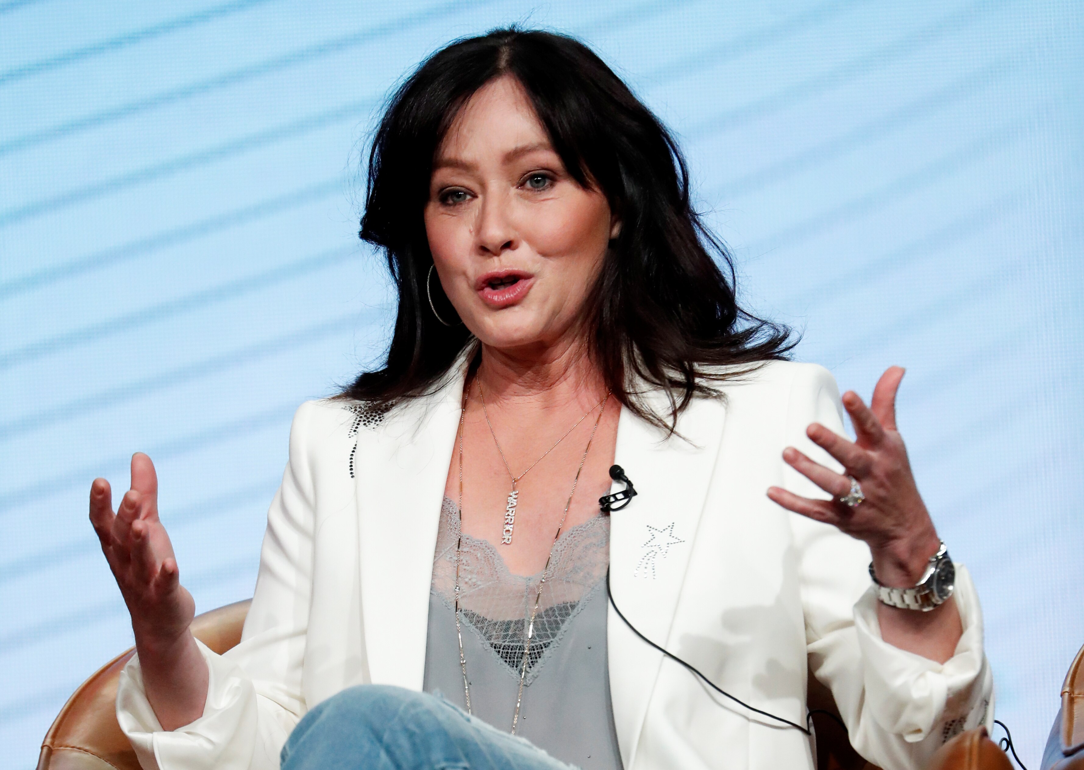 A woman with black hair in a white blazer speaks with her hands.