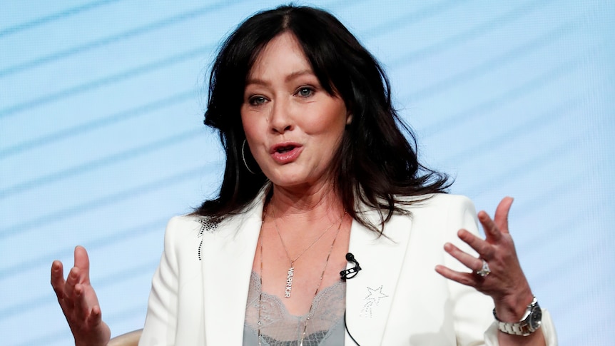 A woman with black hair in a white blazer speaks with her hands.