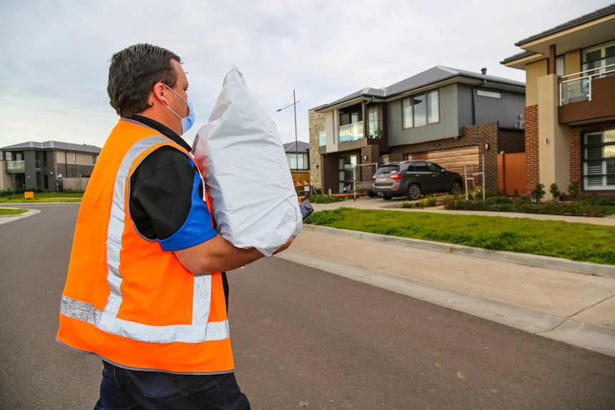 A man in an orange high-vis vest carries a large package as he crosses a suburban street.