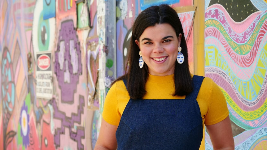 A woman with straight, dark hair, wearing a yellow top and blue overalls, in front of a mural.