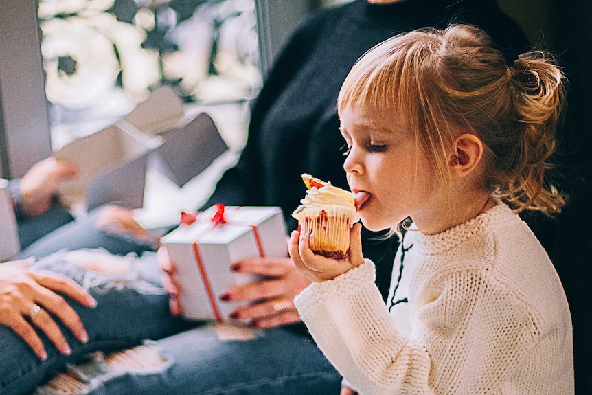 A young girl wearing a white jumper licks the icing off a cupcake while people hold presents and sit with her.