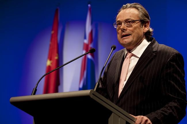 Geoff Raby, with grey hair and glasses, stands behind a lectern, speaking, with the Chinese and Australian flags behind him.