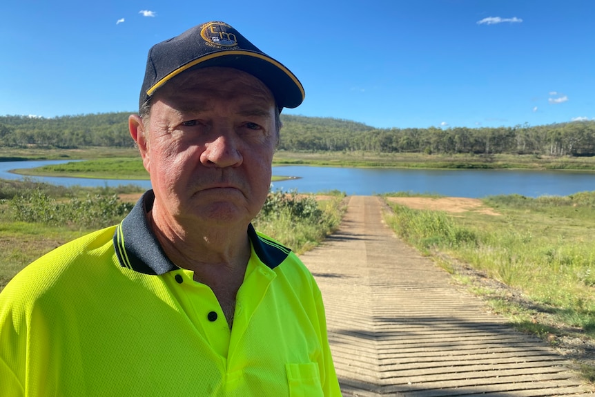 A frowning man with a cap and a yellow high-visibility shirt standing outside, with green foliage behind him and a very low dam