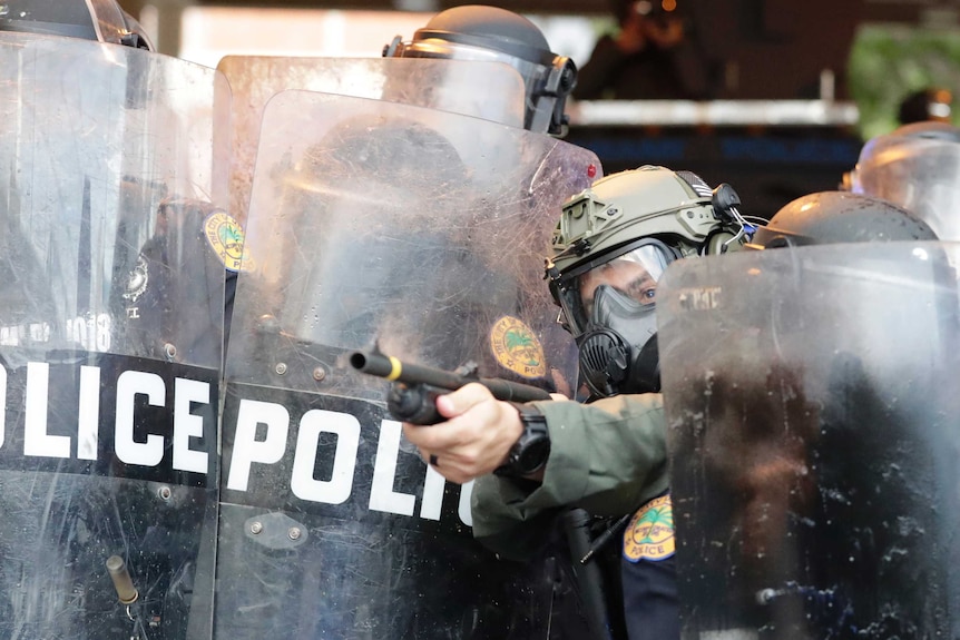 A policeman looks out into a crowd as he prepares to fire rubber bullets. He stands among other officers in riot gear.