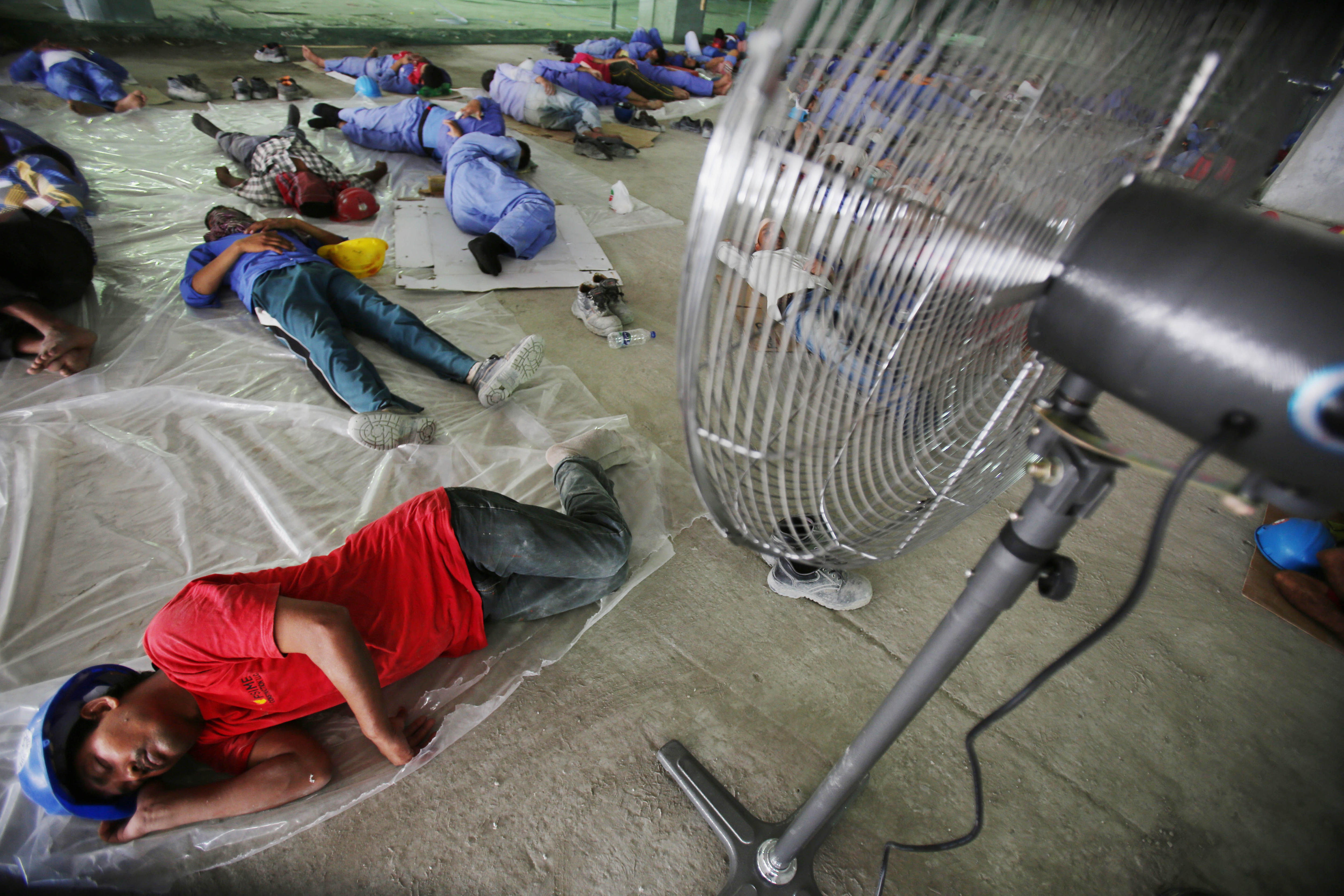 Un grupo de trabajadores duerme sobre un suelo de cemento bajo un ventilador.
