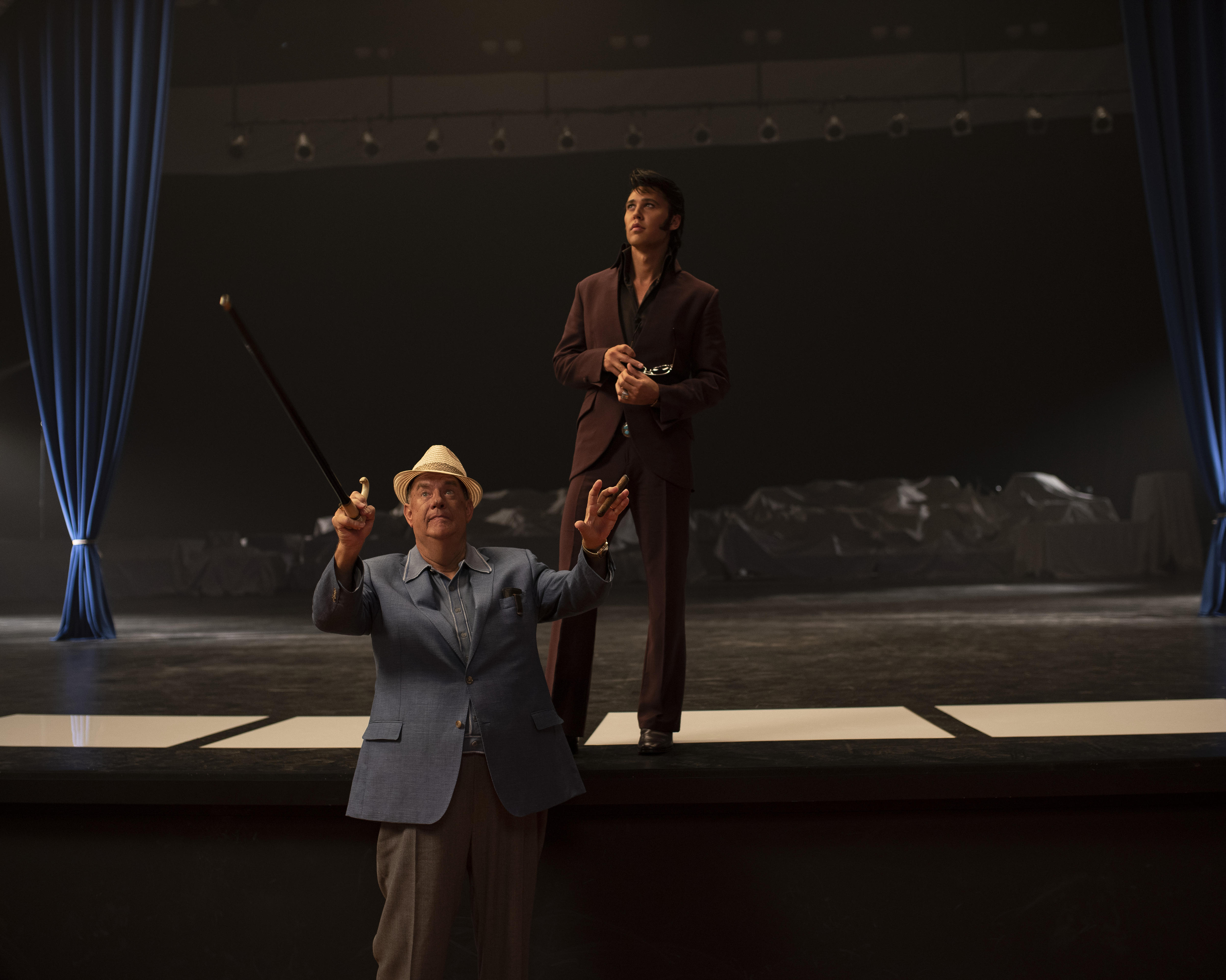 Older white man in grey suit and panama hat holds a walking stick up in front of stage upon which Elvis stands in burgundy suit.