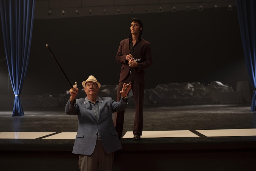 Older white man in grey suit and panama hat holds a walking stick up in front of stage upon which Elvis stands in burgundy suit.