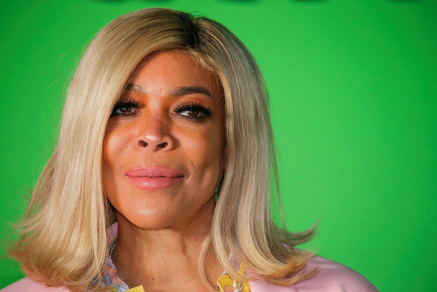 A close up headshot of Wendy Williams with blonde hair against a green background