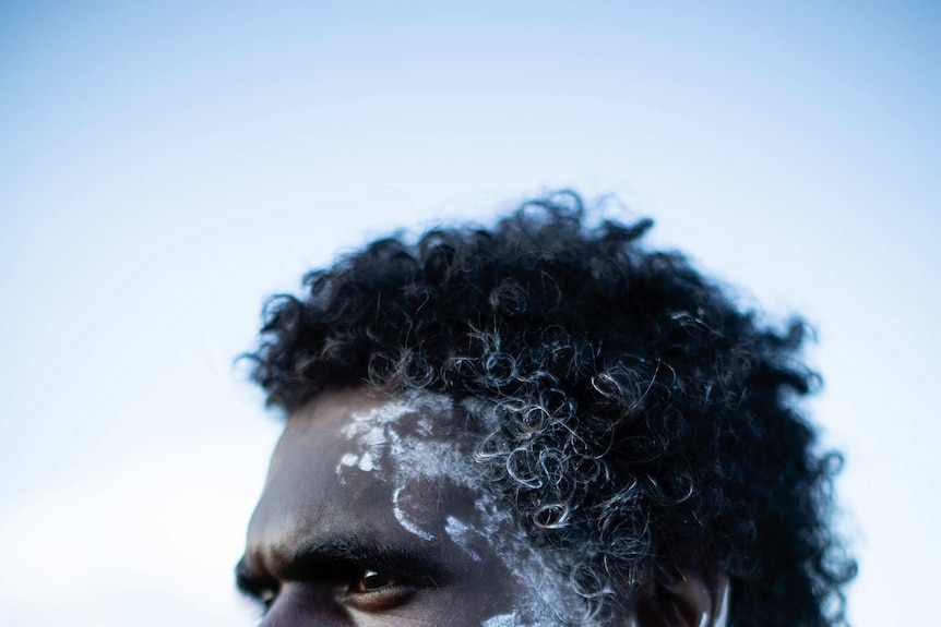Jacob Junior Nayinggul stands in a field in a side profile portrait photo with white paint on the left side of his face.