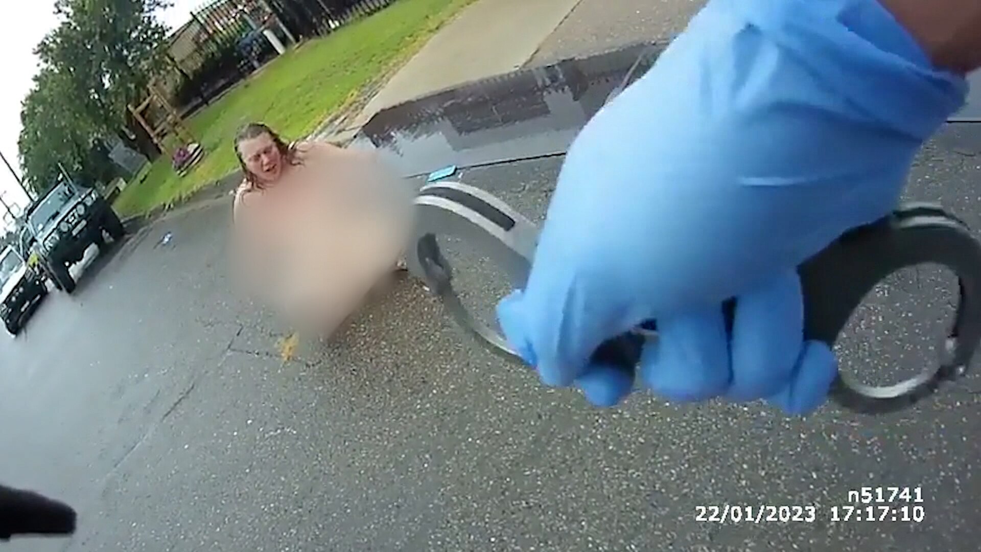 Body-worn camera footage of a naked woman (her body blurred) sits on a street, he face pained. An officer holds handcuffs. 
