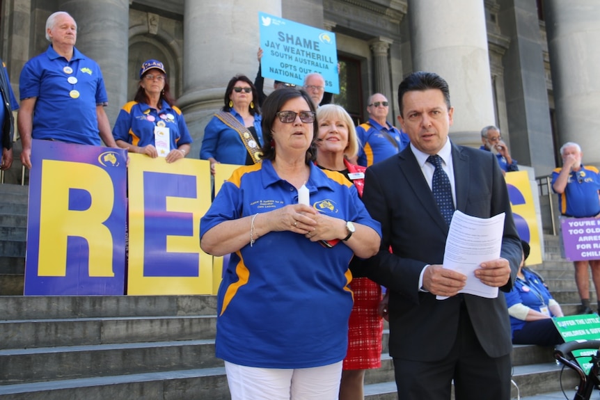 Nick Xenophon joins abuse survivors on the steps of Parliament House, with blue and yellow banners displayed.