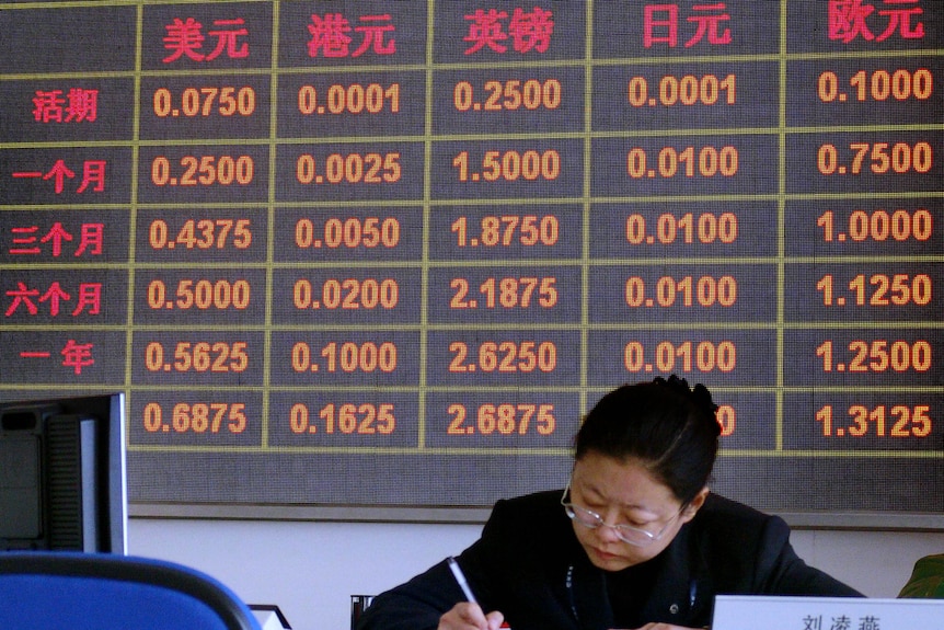 a woman in working in a bank in China, behind her there is a LED screen showing the bank's intersts rate