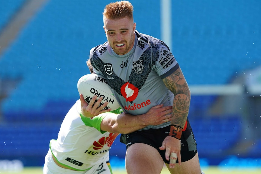 A Warriors NRL player holds the ball with his right arm as he is tackled by a Canberra Raiders opponent.