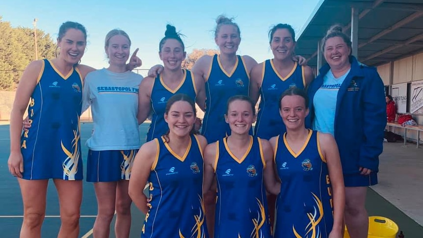 A netball team wearing blue uniforms with yellow trim pose beside a netball court.