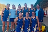 A netball team wearing blue uniforms with yellow trim pose beside a netball court.