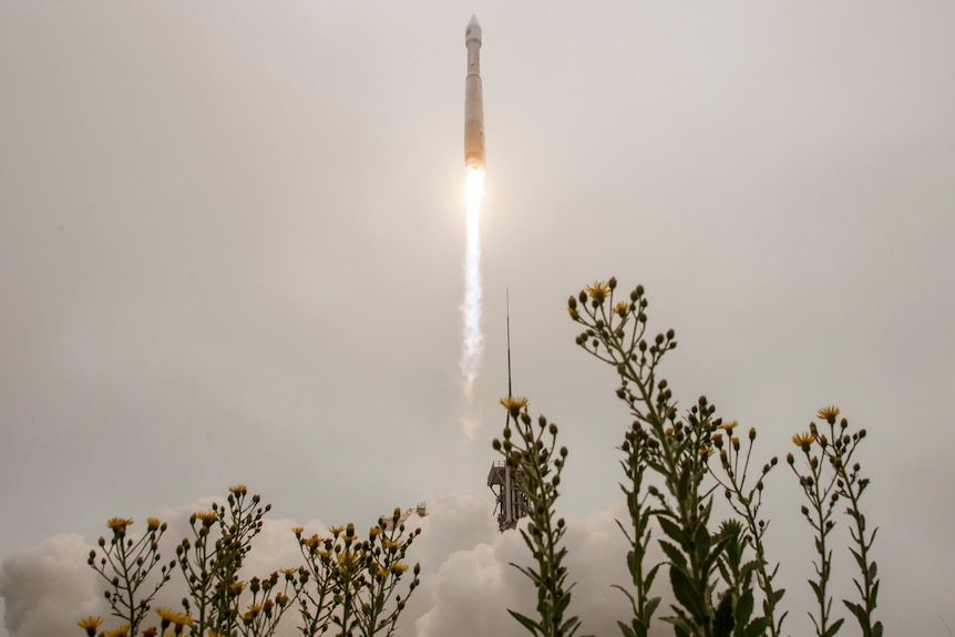 A rocket launches with long blast with flowers and plumes of smoke and fuel below