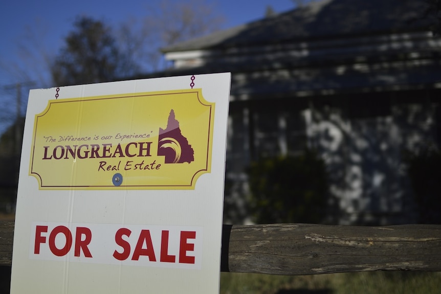 For Sale sign in the foreground with a house in the background.