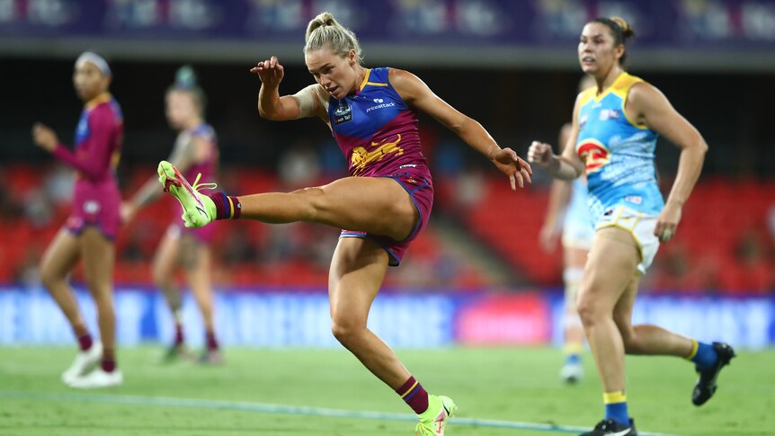 A Brisbane Lions AFLW player kicks through the ball as an opposition player runs behind her during a game..
