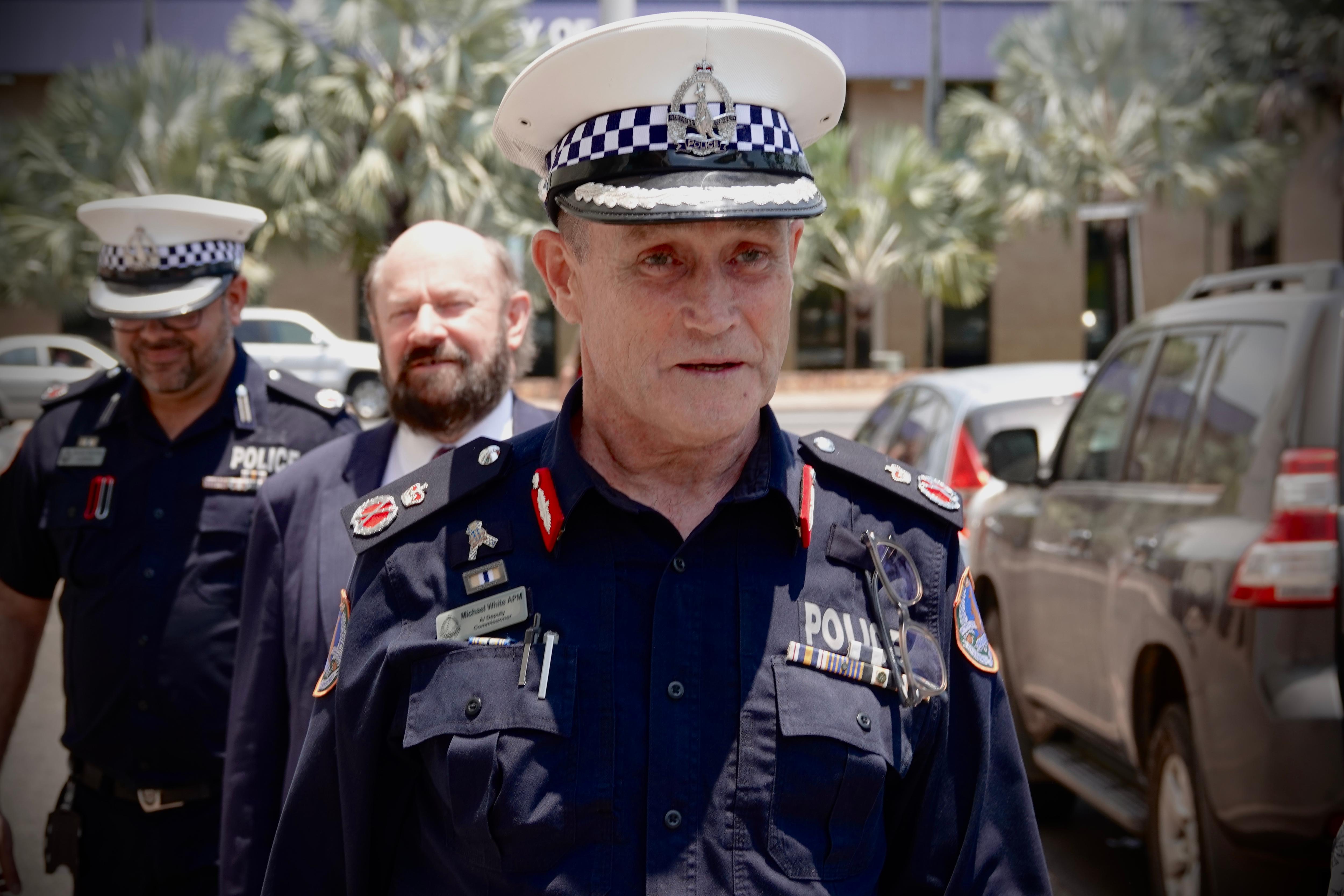 A police officer in the foreground walks along a street. A man in a suit and another police officer walk behind him