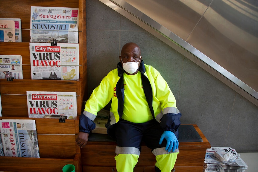 A man in bright hi-vis yellow clothing stares into the camera sitting next to a newspaper stand in front of an escalator.