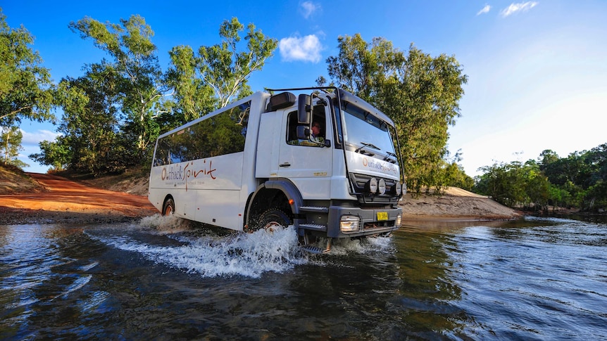 A photo of a bus passing off a dirt road into a shallow creek. The sky is blue.