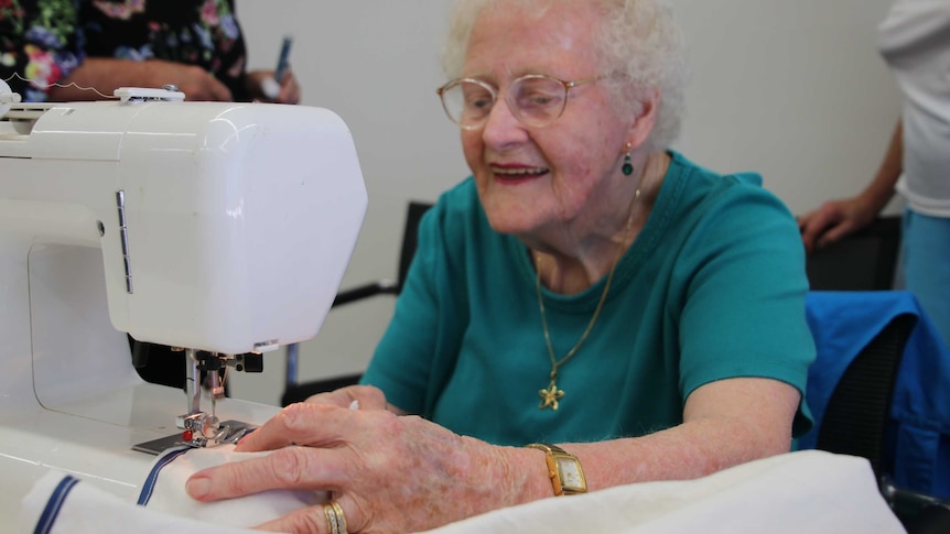 An elderly woman smiles as she uses a sewing machine to stitch the edge of white material.