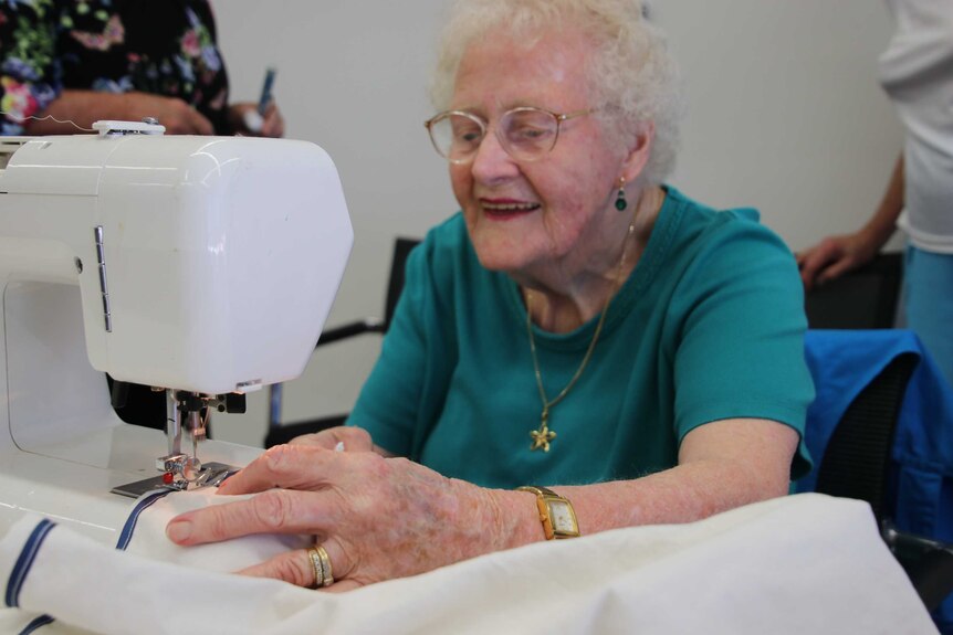 An elderly woman smiles as she uses a sewing machine to stitch the edge of white material.