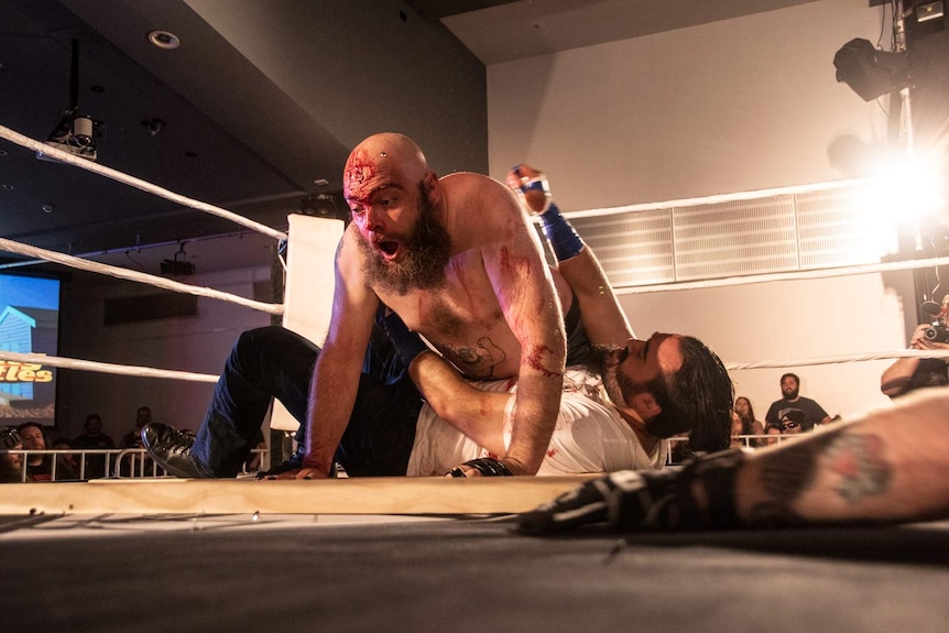 A man bleeding from the forehead and with look of surprise uses his body to pin another man to the floor of a boxing ring.
