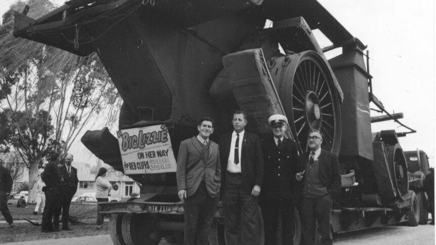 Black and white photo of giant tractor with men standing beside the wheel