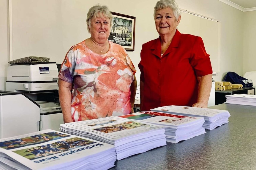 Two women, wearing red and pink stand behind a table in a printing room, on the table are large stacks of newsletters.