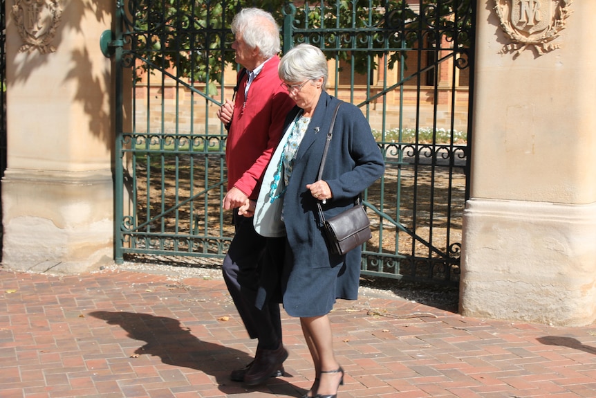 A man and a woman hold hands as they walk in front of an ornate fence.