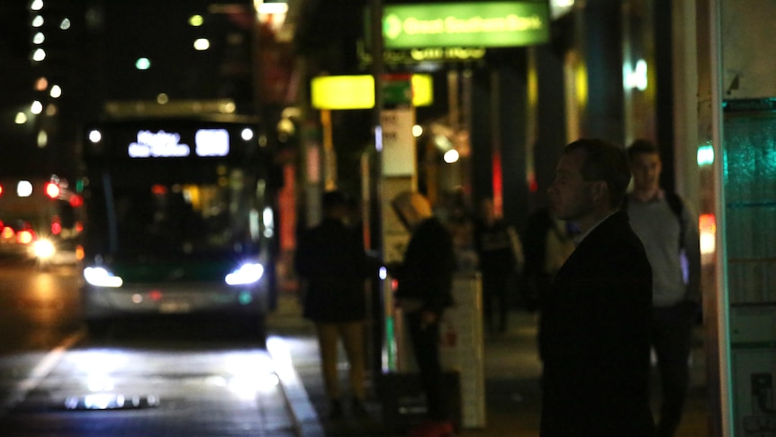 A dark CBD street scene as people wait for buses on William Street as one approaches.