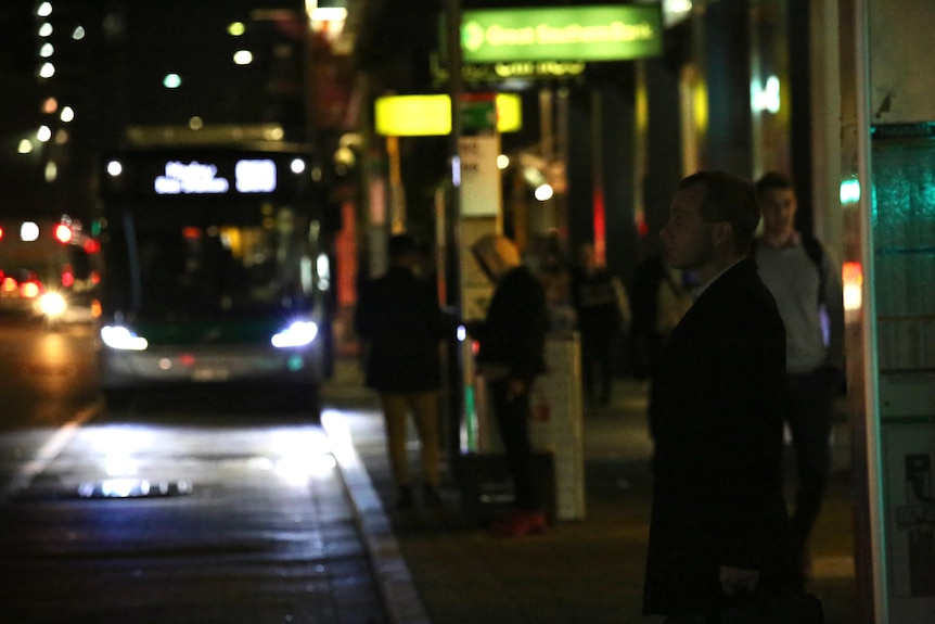 A dark CBD street scene as people wait for buses on William Street as one approaches.