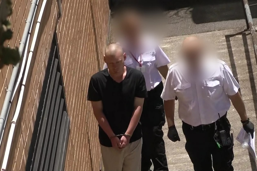 A bald man wearing handcuffs walking with two prison guards. 