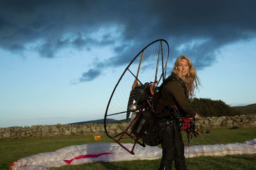 A woman stands with a  fan-like object on her back, in front an unfurled parachute on the ground.