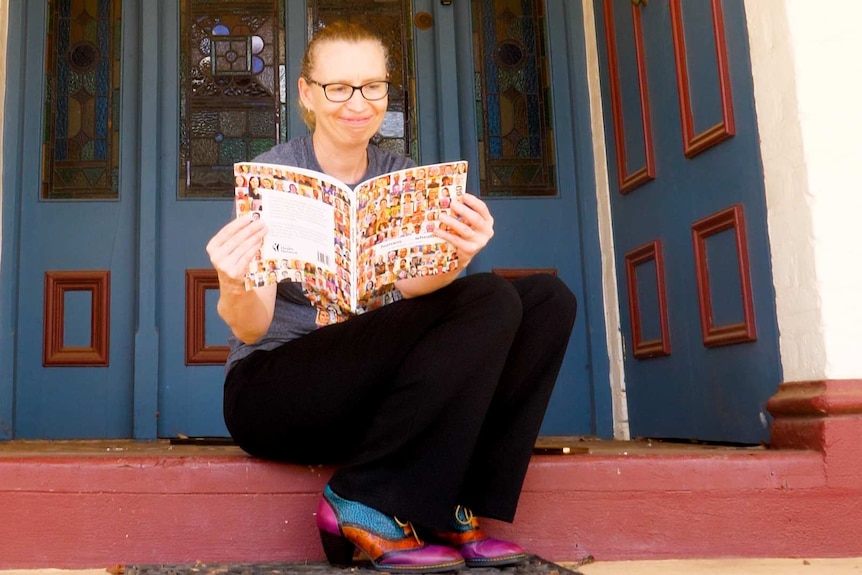 Woman with glasses sitting on a doorstep of her Northam home reading the Humans of the Wheatbelt book.