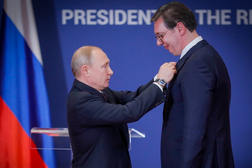 Vladimir Putin reaches out to pin a badge on Aleksandar Vucic's tie. Vucic is significantly taller than Putin