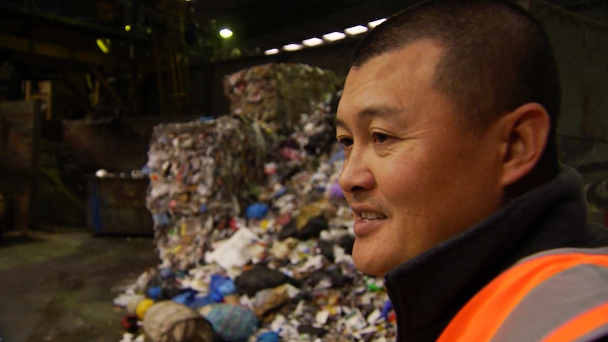 Nathan Ung, wearing an orange high-visibility vest, at a recycling plant. Rubbish is visible behind him.